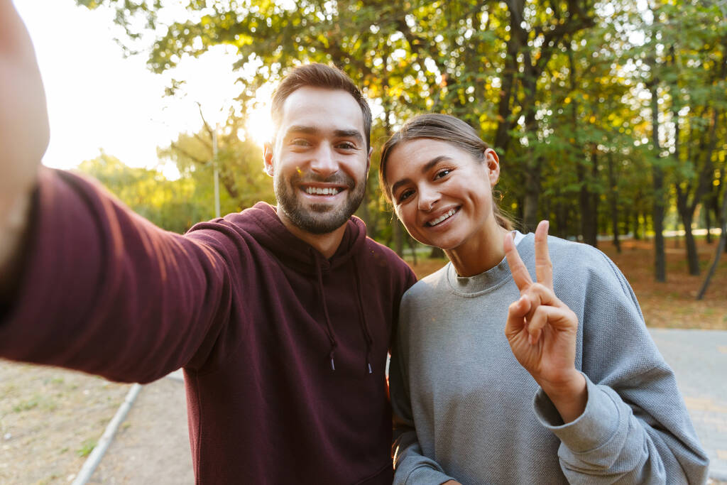 Φωτογραφία του νεαρού ευτυχισμένου ζευγαριού σε αθλητικά ρούχα λαμβάνοντας selfie φωτογραφία και gesturing σύμβολο ειρήνης στο πράσινο πάρκο - Φωτογραφία, εικόνα