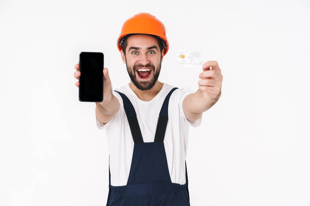 Imagem de alegre otimista jovem construtor positivo no capacete isolado sobre fundo da parede branca segurando cartão de crédito mostrando exibição de telefone móvel
. - Foto, Imagem