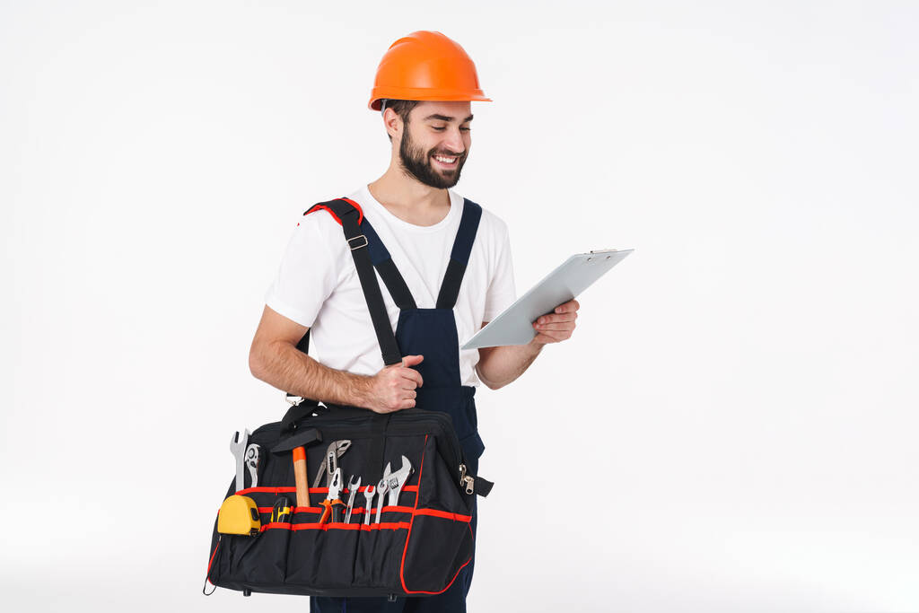 Bild des lächelnden positiven jungen Mannes Bauarbeiter in Helm isoliert über weißen Wand Hintergrund Halterung Tasche mit Ausrüstung Instrumente mit Klemmbrett. - Foto, Bild
