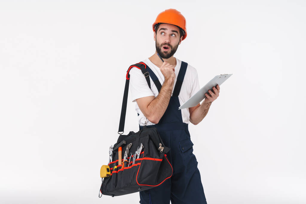 Bild des denkenden jungen Mannes Bauarbeiter in Helm isoliert über weißen Wand Hintergrund Halterung Tasche mit Ausrüstung Instrumente halten Klemmbrett. - Foto, Bild