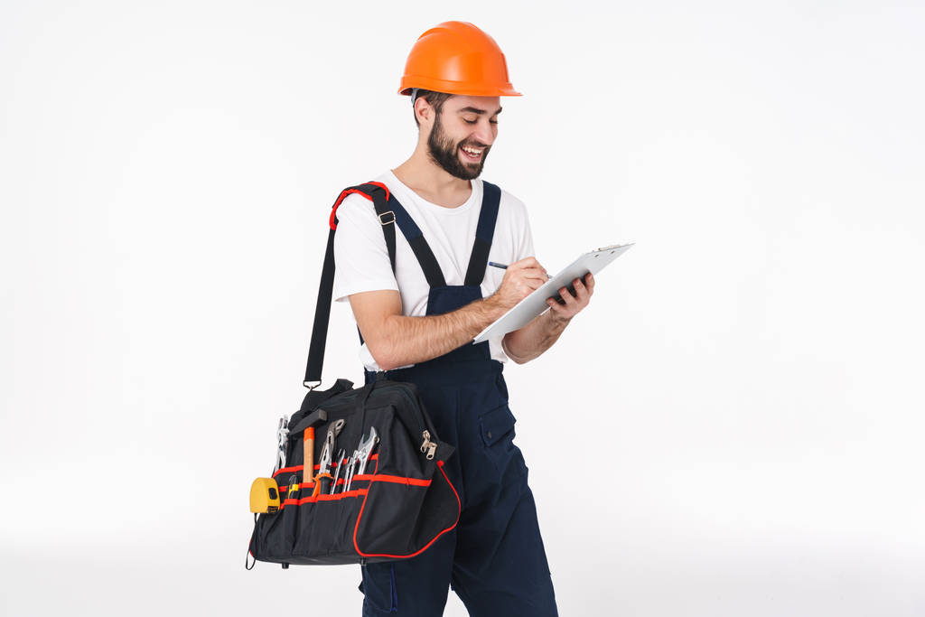 Foto von glücklichen jungen Mann Bauarbeiter in Helm isoliert über weißen Wand Hintergrund halten Tasche mit Ausrüstung Instrumente halten Klemmbrett. - Foto, Bild