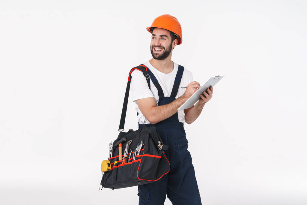Изображение улыбающегося позитивного молодого человека, строителя в шлеме, изолированного на фоне белой стены, держащего сумку с инструментами, держащими планшет
. - Фото, изображение