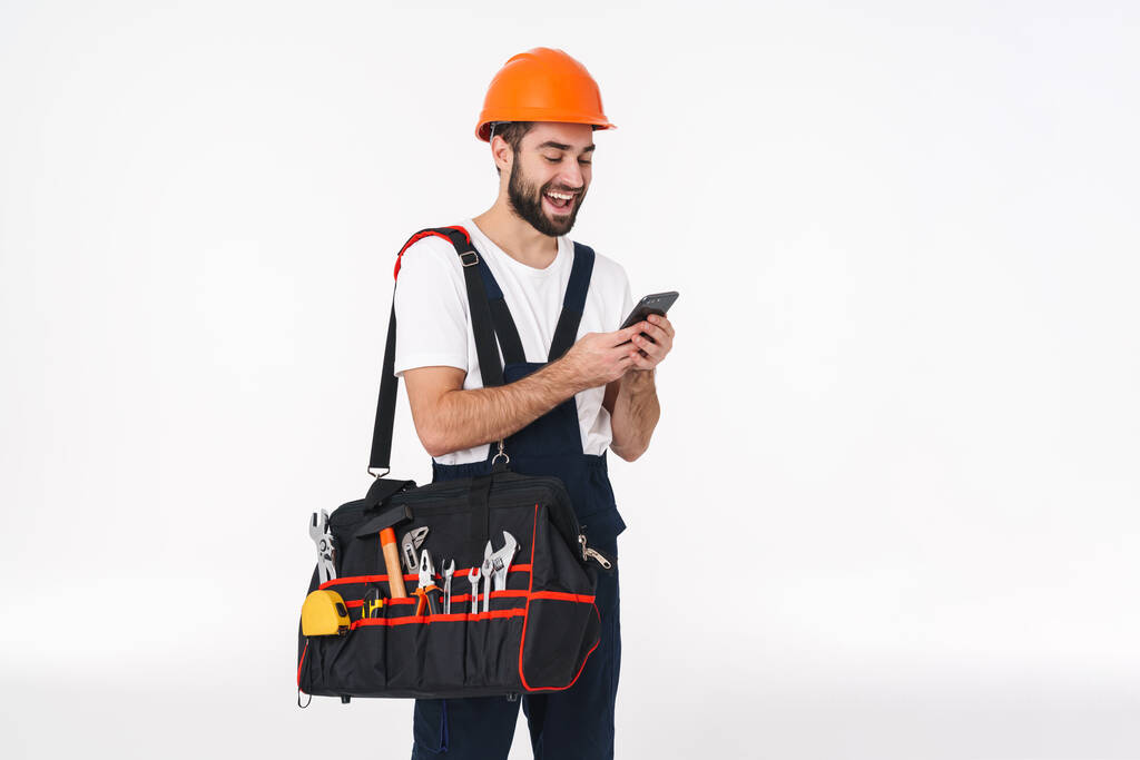 Bild des optimistischen jungen Mannes Bauarbeiter in Helm isoliert über weißen Wand Hintergrund halten Tasche mit Ausrüstung Instrumente mit Mobiltelefon. - Foto, Bild