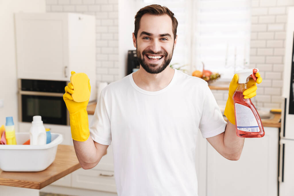 Image de jeune homme positif souriant gai mari au foyer à l'intérieur tenant éponge et spray nettoyant
. - Photo, image