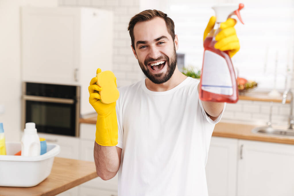 Image of young optimistic positive man househusband indoors holding sponge and cleansing spray. - Photo, Image