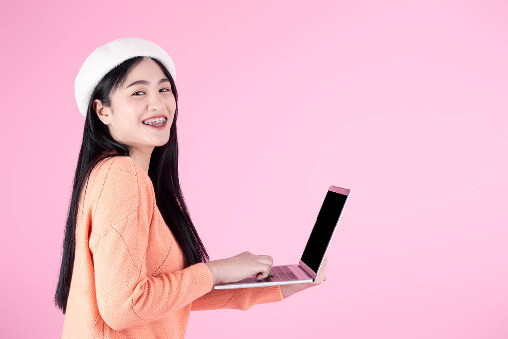 Ευτυχισμένο ασιατικό κορίτσι σε πορτοκαλί πουλόβερ χρησιμοποιώντας φορητό υπολογιστή με αντίγραφο χώρου σε ροζ φόντο. - Φωτογραφία, εικόνα