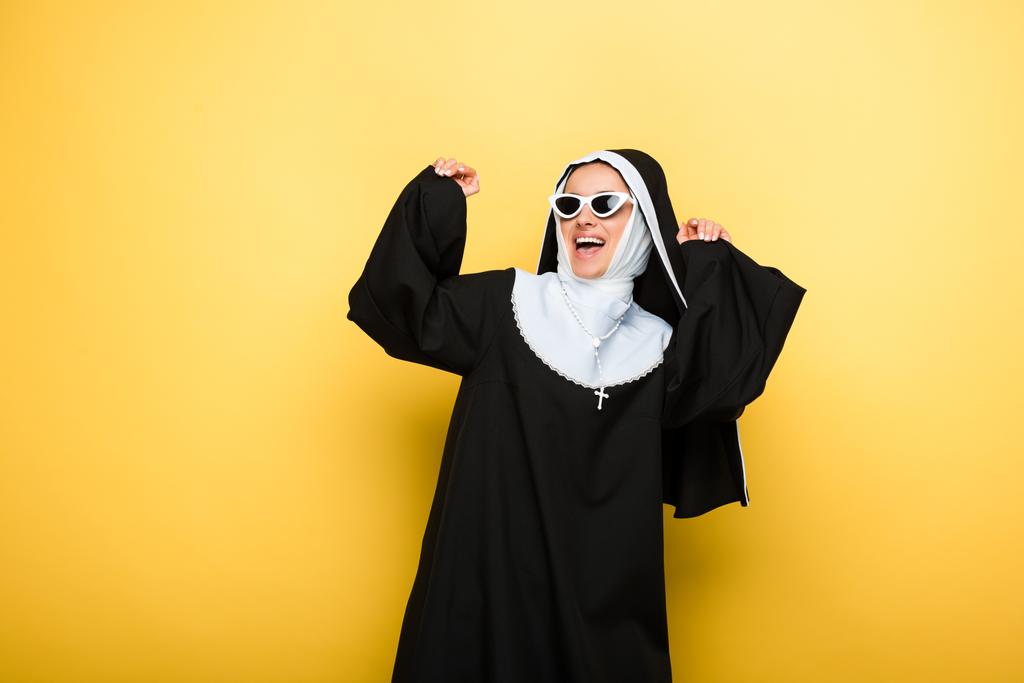 freira emocional feliz atraente dançando em óculos de sol no amarelo
 - Foto, Imagem