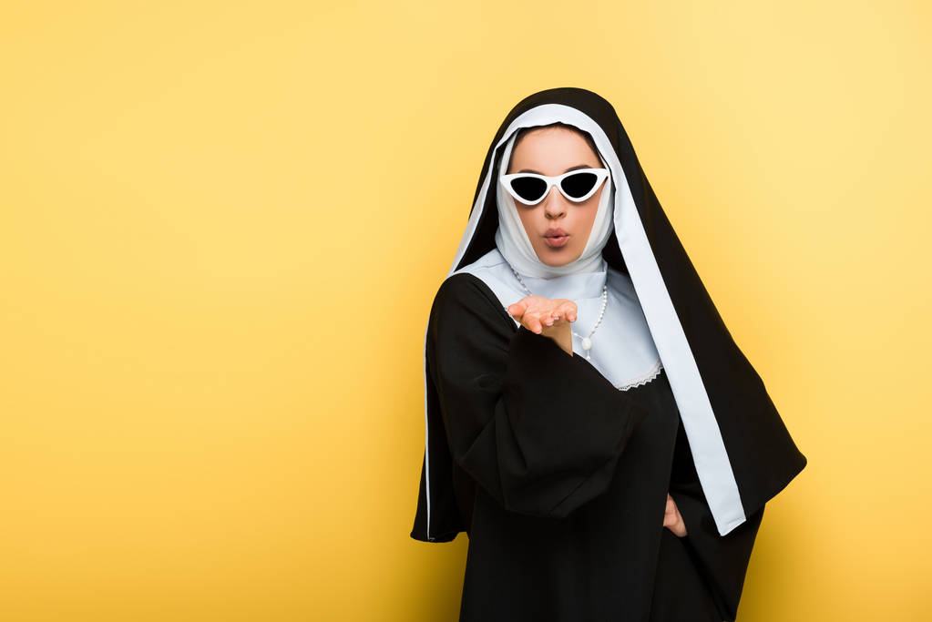 freira atraente em óculos de sol soprando beijo de ar no amarelo
 - Foto, Imagem