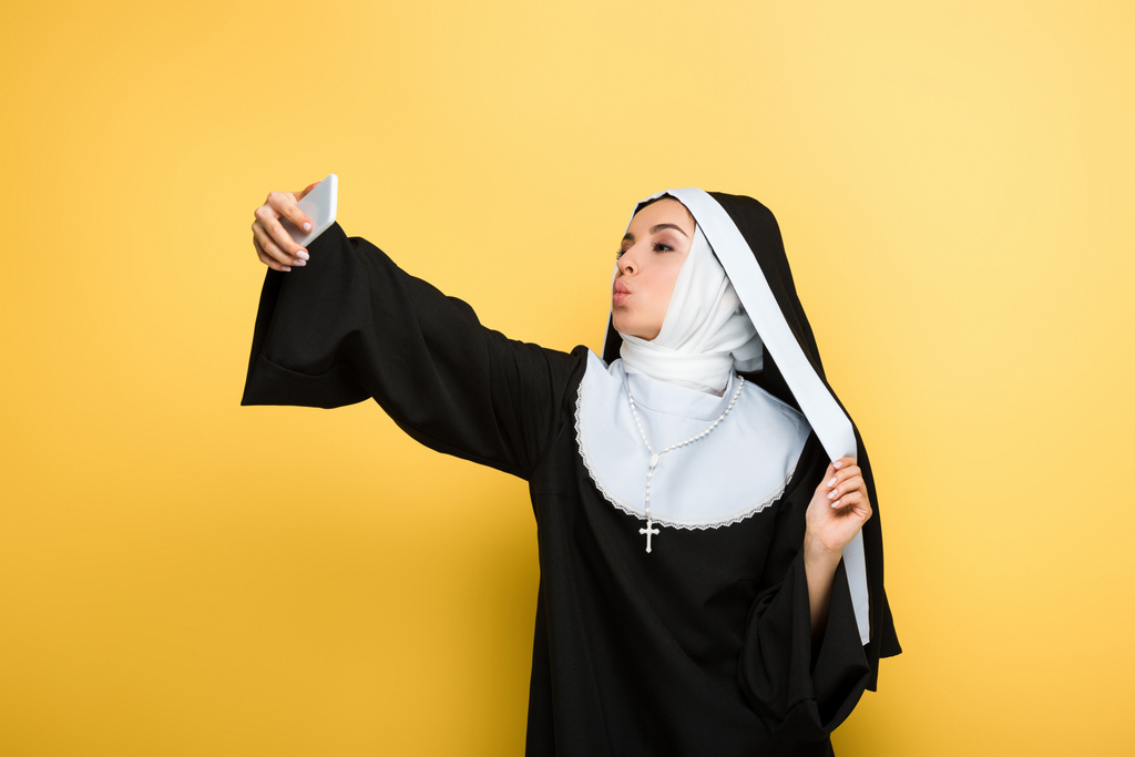 hermosa monja con aire beso tomando selfie en teléfono inteligente aislado en amarillo
 - Foto, imagen