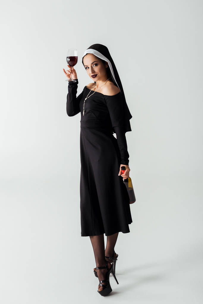 ελκυστική σέξι καλόγρια κρατώντας γυαλί και μπουκάλι κρασί σε γκρι - Φωτογραφία, εικόνα