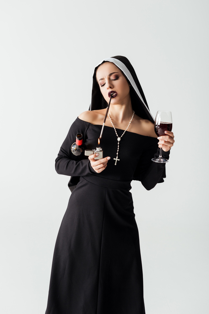 schöne sinnliche Nonne mit Feuerzeug und Wein Rauchen einer Zigarette im Mundstück isoliert auf grau  - Foto, Bild