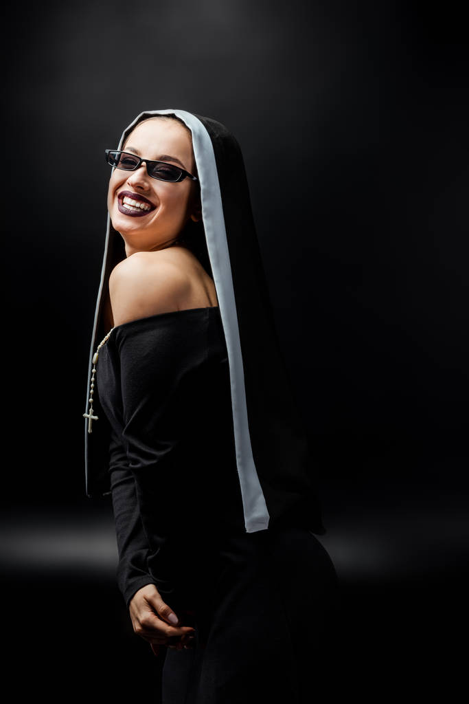 sexy cheerful nun in black dress and sunglasses, on black   - Photo, Image