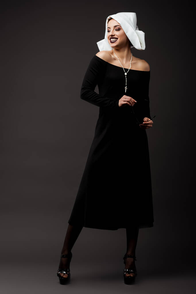 smiling seductive nun in black dress holding sunglasses, on grey   - Photo, Image