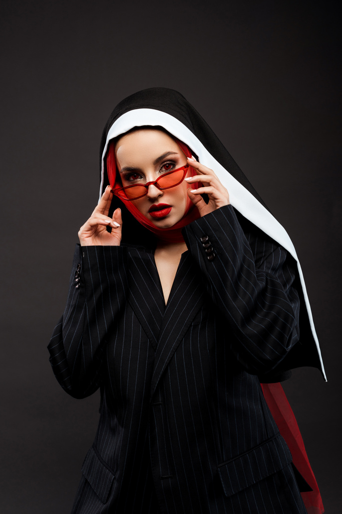 μοντέρνα σέξι δαιμονική καλόγρια με κόκκινα μάτια που ποζάρουν σε μαύρο κοστούμι, κόκκινα γυαλιά ηλίου και μαντήλι, απομονωμένη σε γκρι  - Φωτογραφία, εικόνα