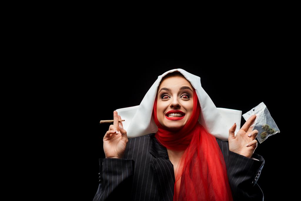 excited demoniac nun holding marijuana joint and buds isolated on black - Photo, Image