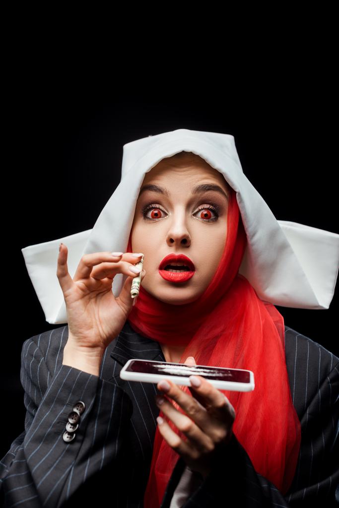 zszokowana demoniczna zakonnica trzymająca banknot dolarowy i smartfona z odizolowanymi na czarno liniami kokainy - Zdjęcie, obraz