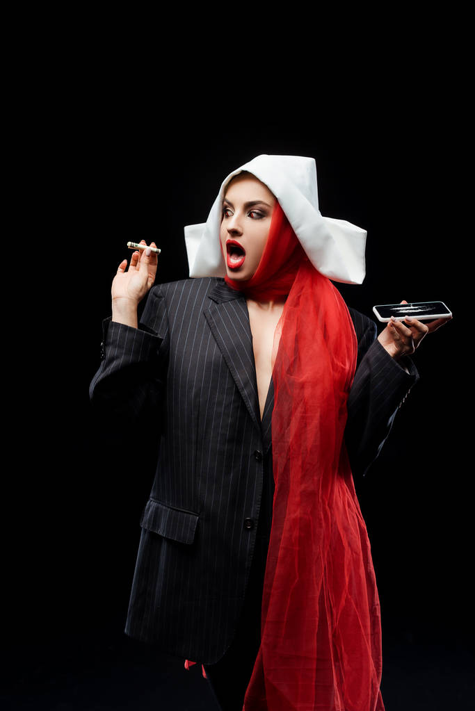 shocked sexy demonic nun holding dollar banknote and smartphone with cocaine lines isolated on black - Photo, Image