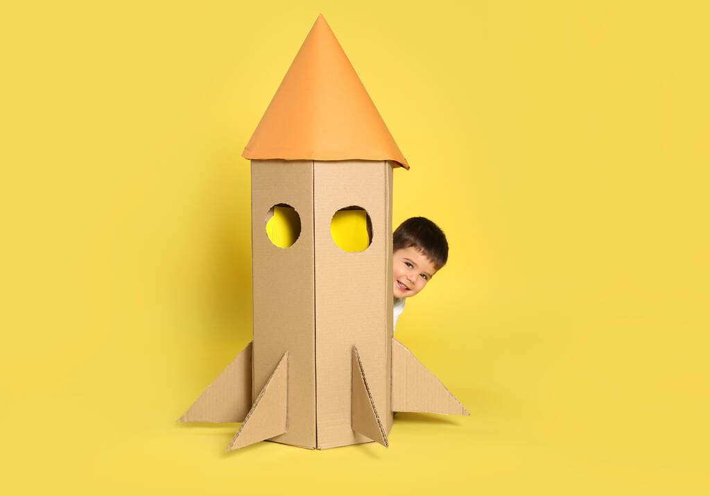 Słodkie małe dziecko bawiące się kartonową rakietą na żółtym tle - Zdjęcie, obraz
