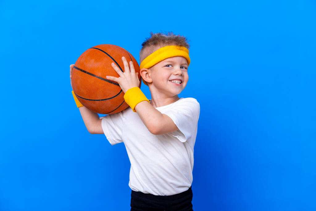 Спортивный мальчик с баскетбольным мячом на фоне синей студии. Тренажерный зал. Детский спортсмен. Деятельность. Спорт. Фитнес, здоровье и энергия. Успех
 - Фото, изображение
