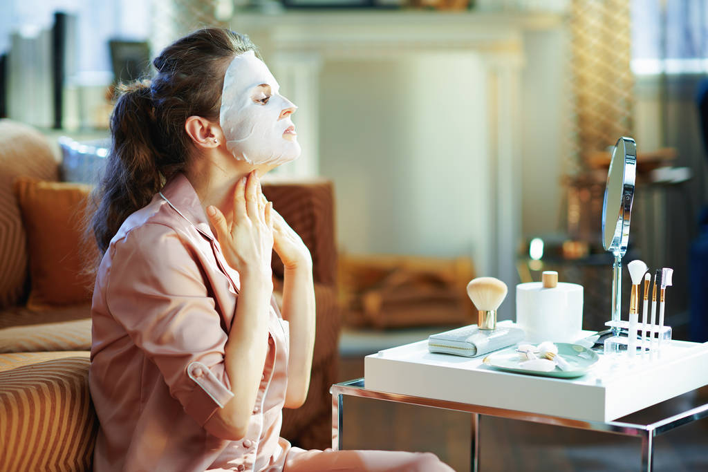 femme d'âge moyen en pyjama à la maison moderne dans la journée ensoleillée d'hiver en appliquant un masque facial feuille blanche et en regardant dans le miroir
. - Photo, image