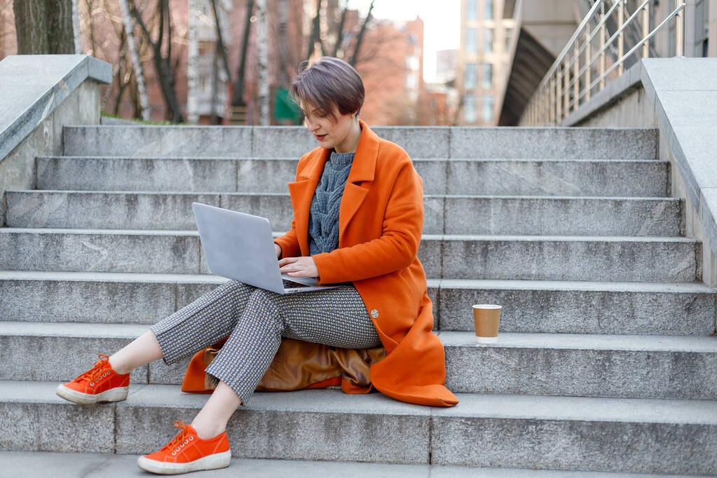 Femme élégante en manteau orange vif dans une scène de ville, en utilisant un ordinateur portable et de travailler dessus pendant une pause. Technologie de plein air. boire du café, thé, cacao
 - Photo, image