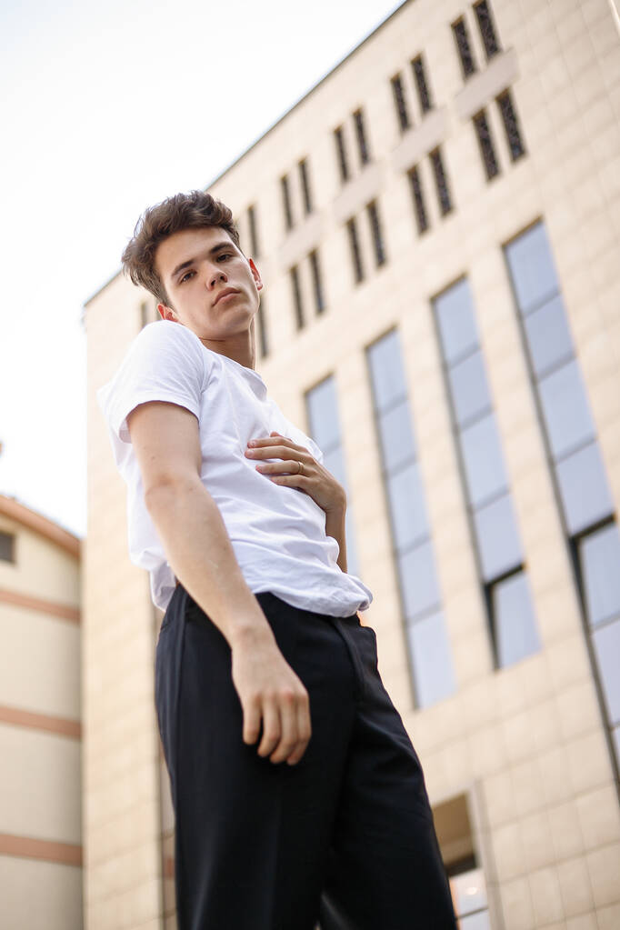 Elegant young man in a fashionable black shirt in a white stylish t-shirt in black pants with a trendy hairstyle rests near a modern business center. Attractive guy in the street on a summer day. - Photo, Image