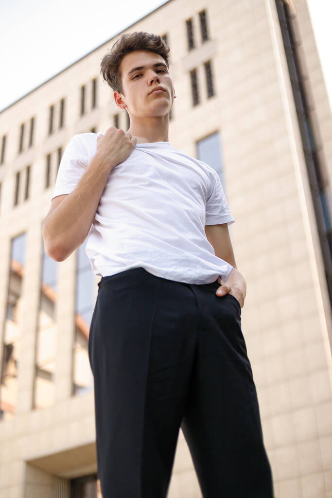 Элегантный молодой человек в модной черной рубашке в белой стильной футболке в черных штанах с модной прической находится рядом с современным бизнес-центром. Привлекательный парень на улице в летний день
. - Фото, изображение