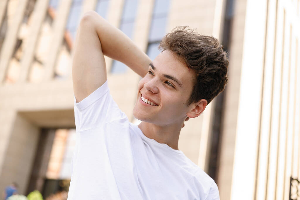 Eleganter junger Mann lächelt in einem modischen schwarzen Hemd in einem weißen, stylischen T-Shirt in schwarzer Hose mit trendiger Frisur in der Nähe eines modernen Business Centers. Porträt aus nächster Nähe - Foto, Bild