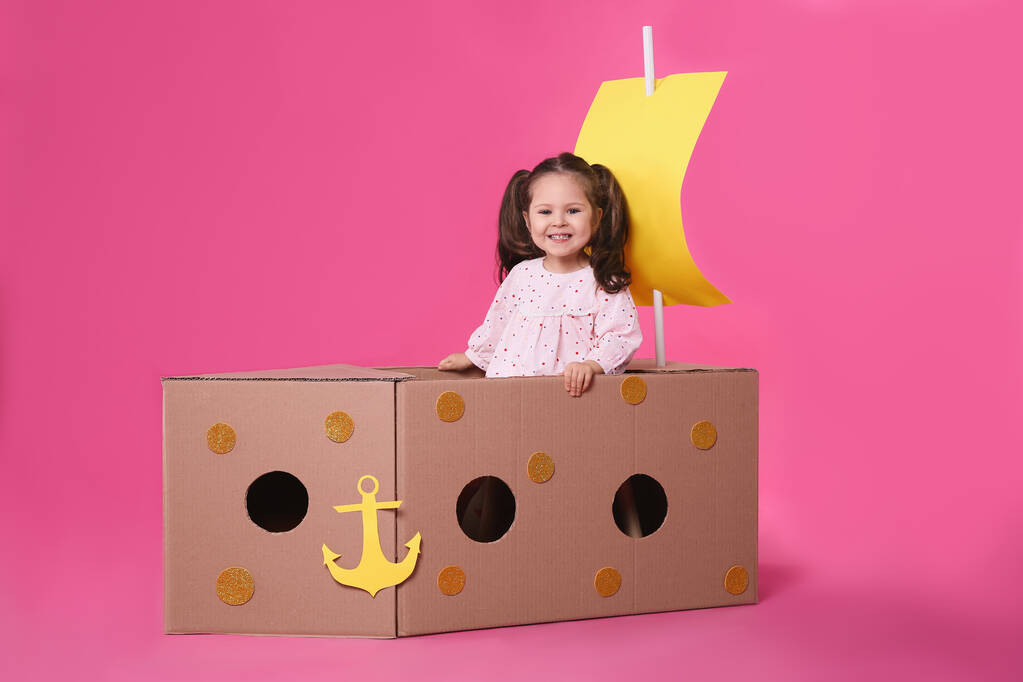 Klein kind spelen met schip gemaakt van karton doos op roze achtergrond - Foto, afbeelding