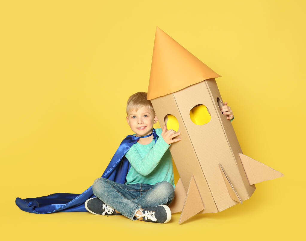 Niño pequeño en capa jugando con cohete hecho de caja de cartón sobre fondo amarillo
 - Foto, imagen