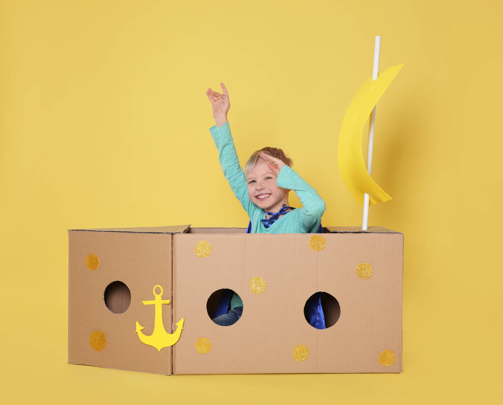 Little child playing with ship made of cardboard box on yellow background - Photo, Image