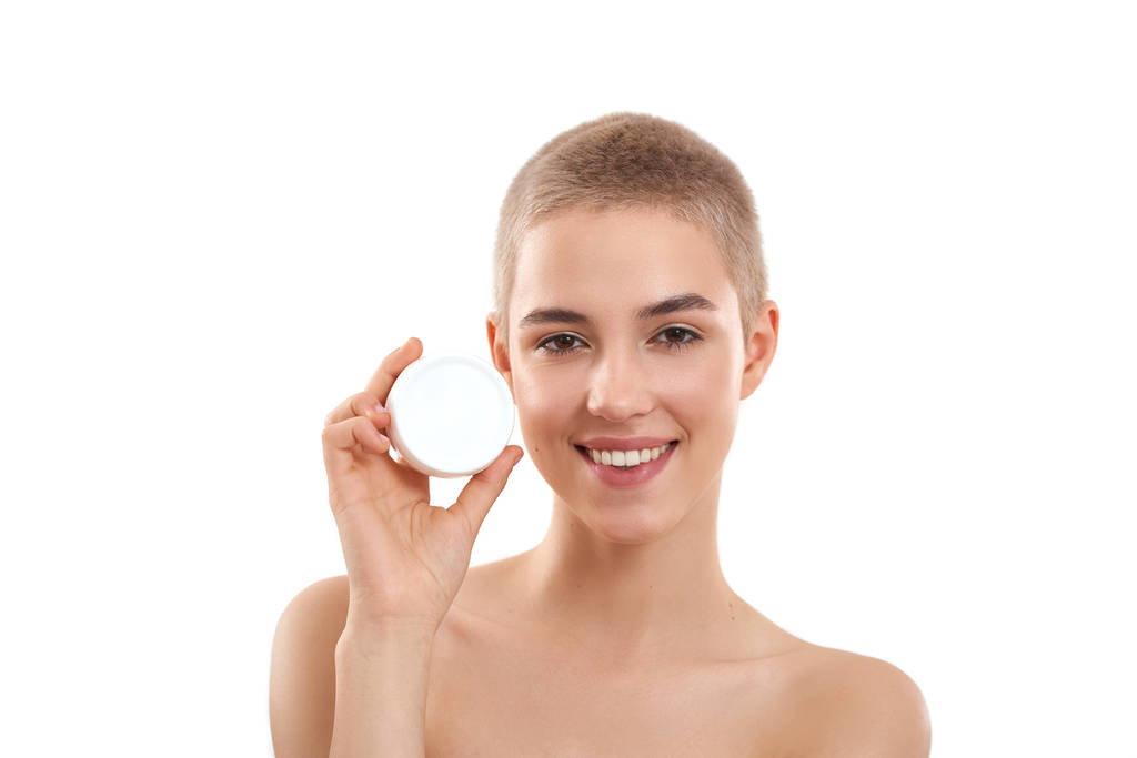 Perfect face cream. Young attractive blonde woman with short haircut demonstrating cosmetic product and smiling at camera while standing in studio against white background - 写真・画像