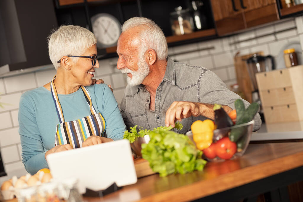 Happy Senior couple cooking healthy meal together in home kitchen digital tablet helps them to prepare food. - Foto, Imagen