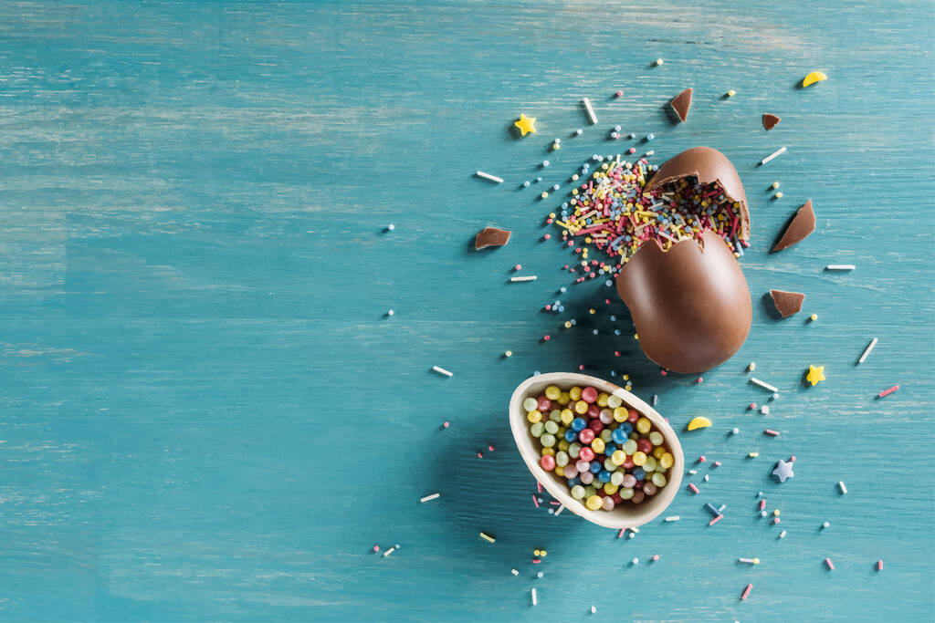 верхний вид шоколадного яйца с пасхальными брызгами на голубом деревянном столе
 - Вектор,изображение