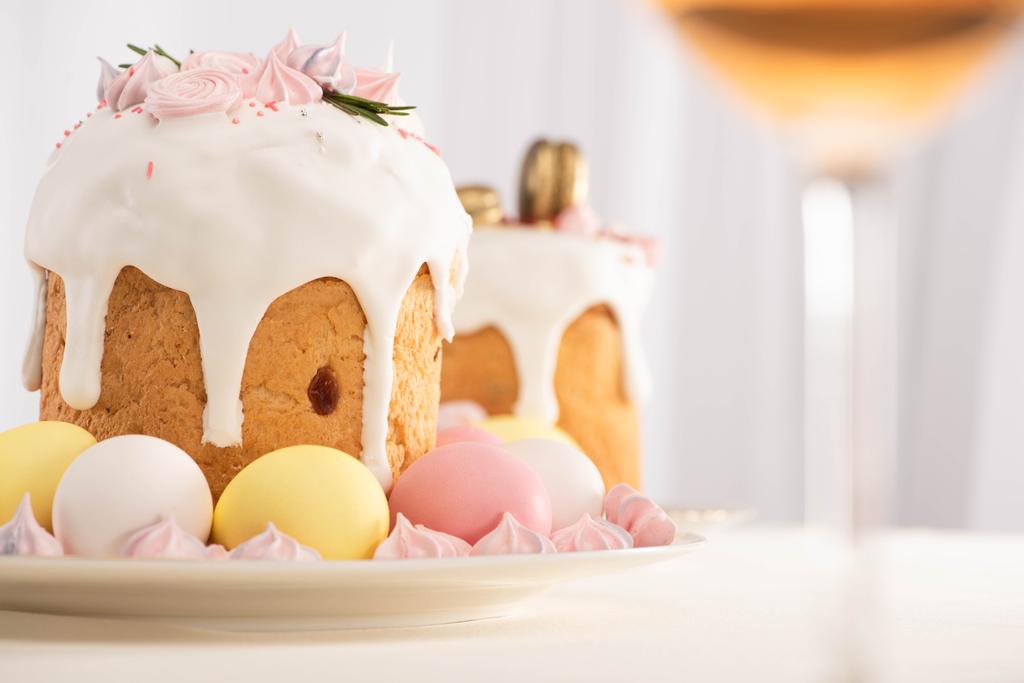 selective focus of delicious Easter cakes decorated with meringue near colorful eggs on plates and wine glass - Photo, Image