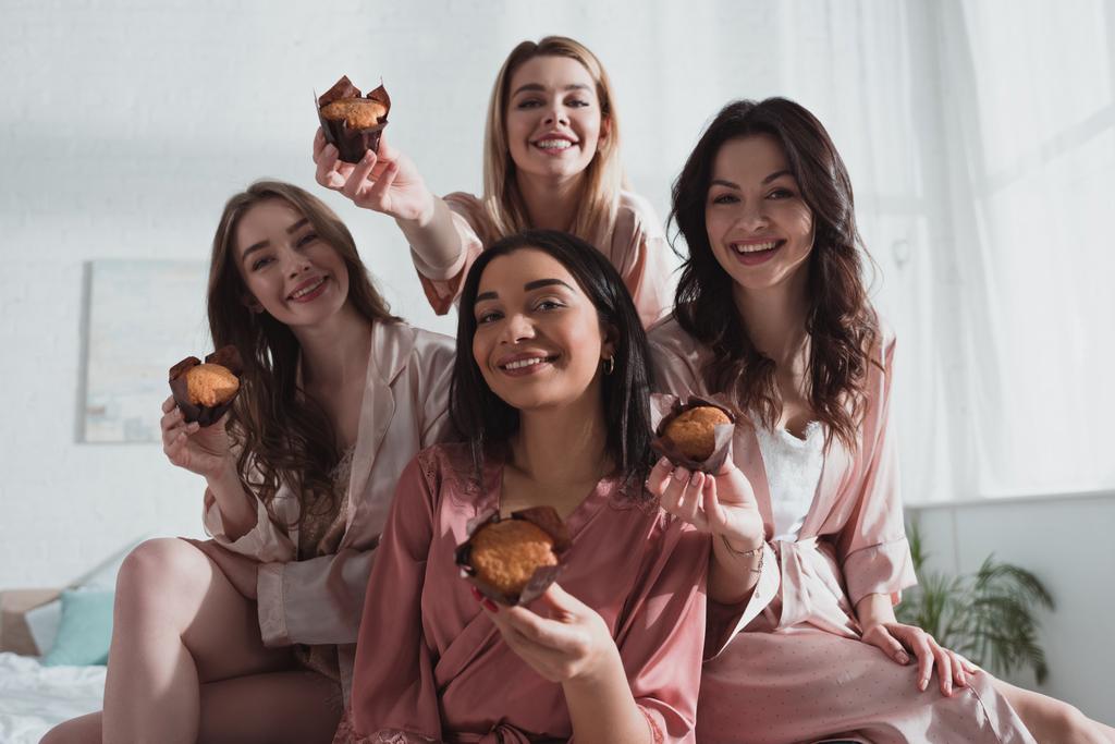 Selektiver Fokus multikultureller Frauen, die lächeln und Muffins auf der Junggesellenabschiedsparty zeigen - Foto, Bild