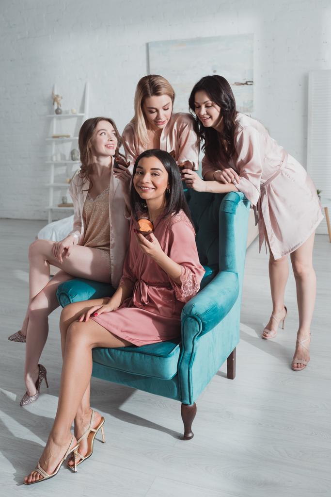 Selective focus of multiethnic women with muffins happy together in armchair in room at bachelorette party - Photo, Image