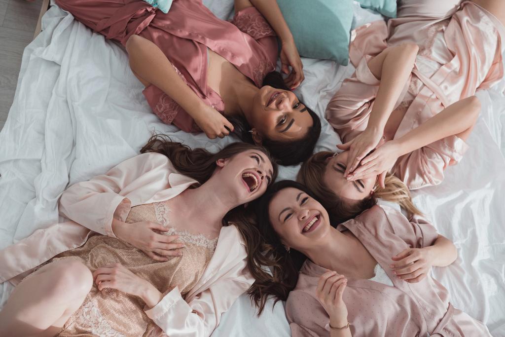 Вид сверху на счастливых мультикультурных женщин, улыбающихся и лежащих на кровати на вечеринке в Лос-Анджелесе
 - Фото, изображение