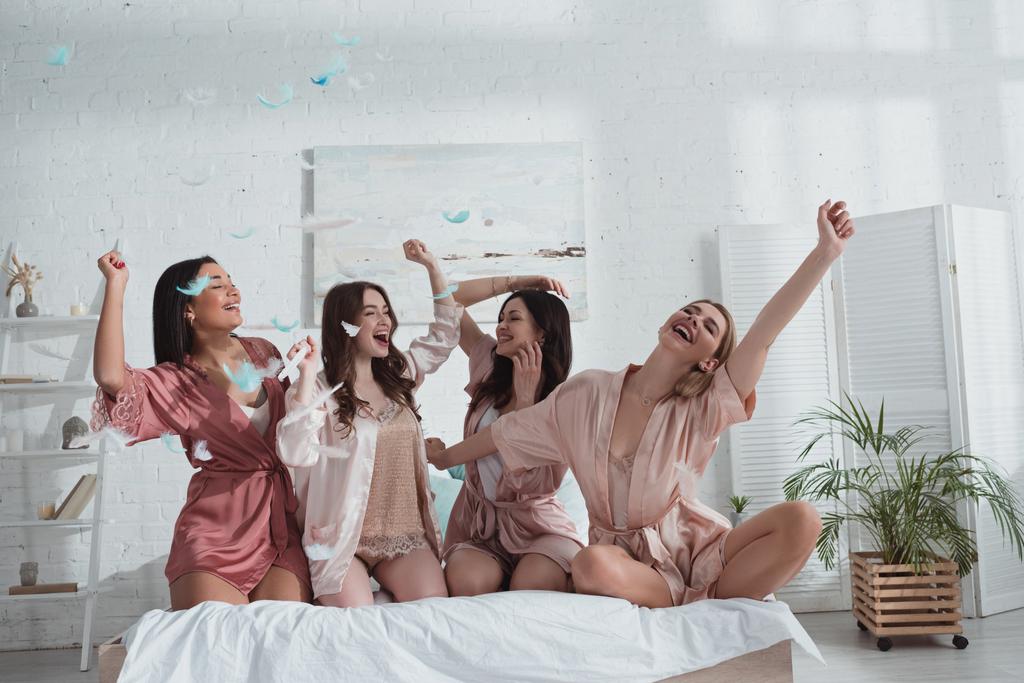 Šťastné a vzrušené multikulturní ženy s rukama ve vzduchu na posteli s peřím v pokoji na rozlučce se svobodou - Fotografie, Obrázek