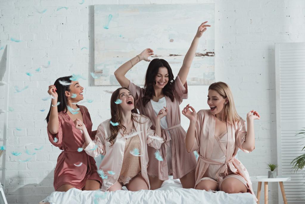 Szczęśliwe i podekscytowane wielonarodowe kobiety z rękami w powietrzu na łóżku z piórami w pokoju na wieczorze panieńskim - Zdjęcie, obraz