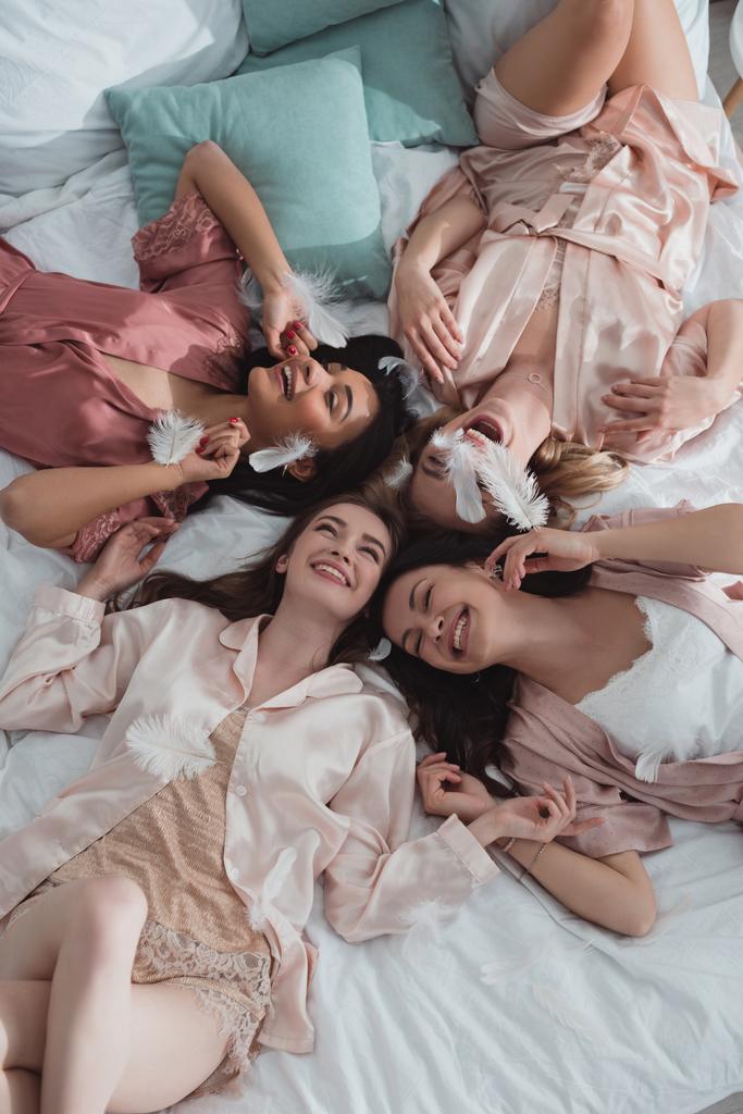Bekarlığa veda partisinde tüylerle yatakta dinlenen mutlu ve heyecanlı çok ırklı kadınların en iyi görüntüsü. - Fotoğraf, Görsel