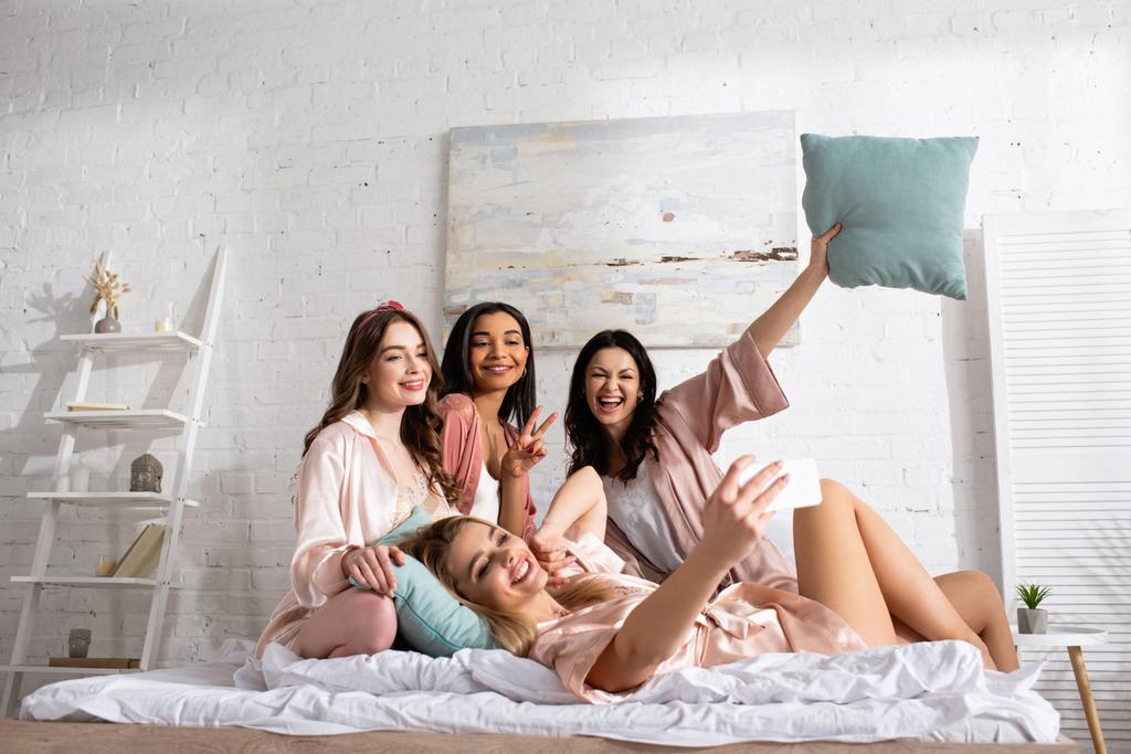 Happy multiethnic women with pillows taking selfie on bed at bachelorette party - Photo, Image
