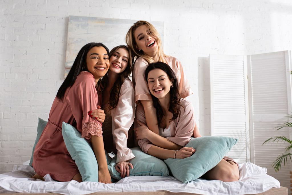 Šťastní multietničtí přátelé dívající se na kameru a usmívající se spolu s polštáři na posteli na rozlučce se svobodou - Fotografie, Obrázek