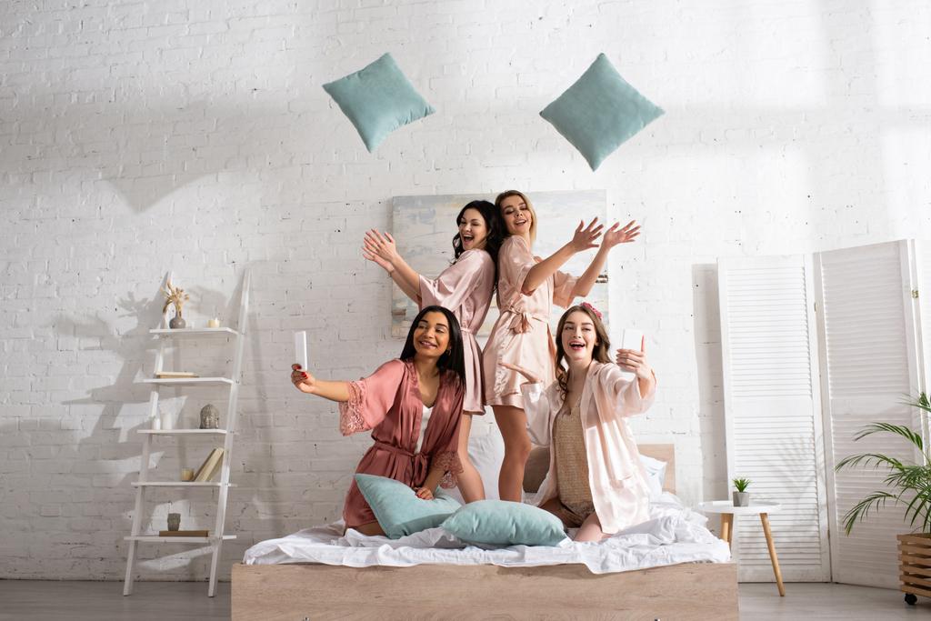 Glückliche und positive multikulturelle Frauen lächeln, werfen Kissen hoch und machen Selfie mit Smartphones im Bett  - Foto, Bild