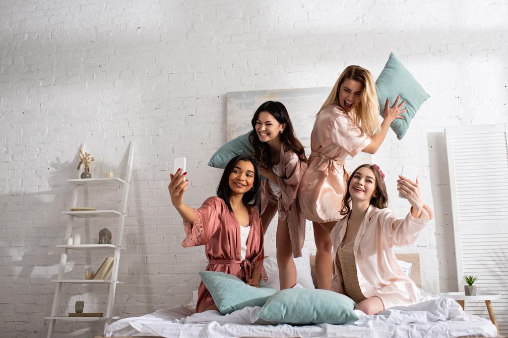 Szczęśliwe wielokulturowe kobiety uśmiechnięte, trzymające poduszki i robiące selfie ze smartfonami na łóżku na wieczorze panieńskim - Zdjęcie, obraz