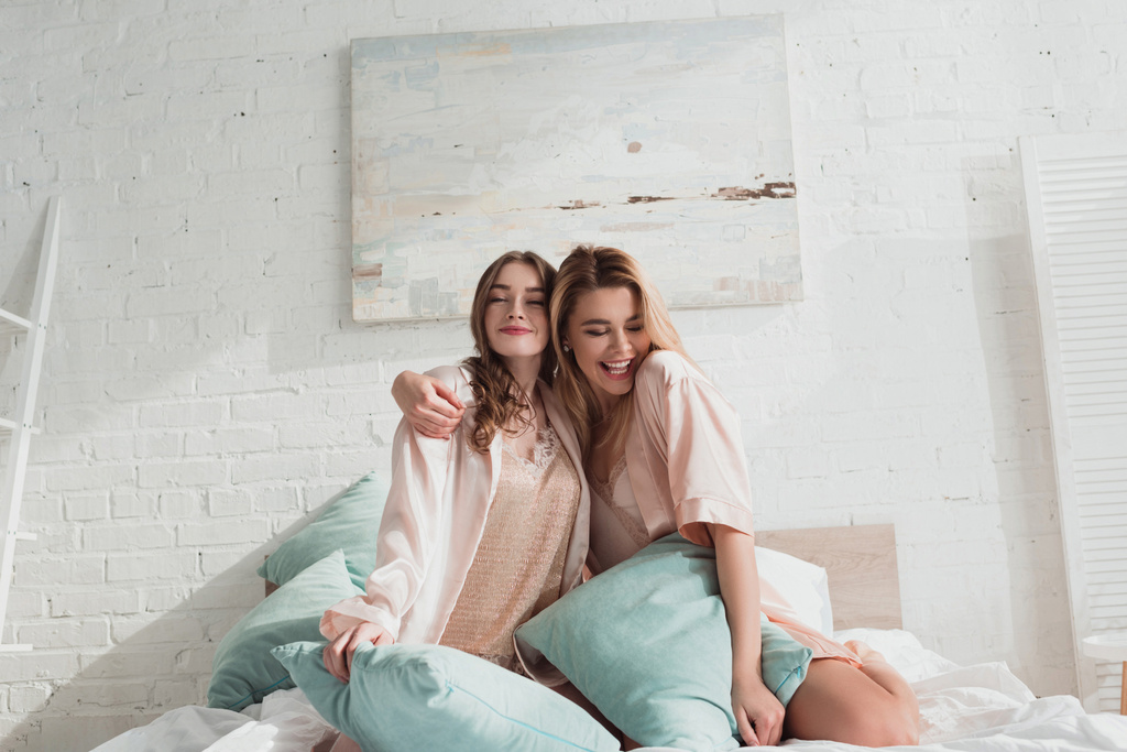Mujeres atractivas abrazándose y sonriendo con almohadas en la cama en la despedida de soltera
 - Foto, imagen