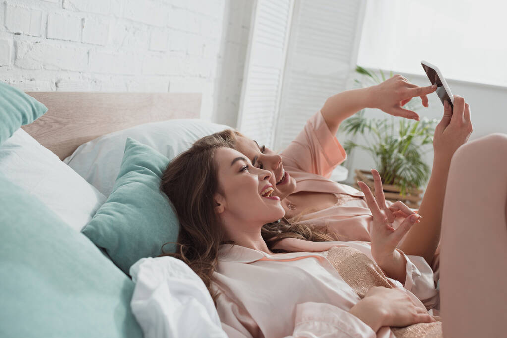 Podekscytowane i szczęśliwe kobiety robiące selfie i odpoczywające na łóżku na wieczorze panieńskim - Zdjęcie, obraz