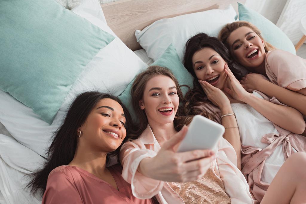Υψηλής γωνίας άποψη του ενθουσιασμένοι πολυπολιτισμικές γυναίκες λήψη selfie ενώ στηρίζεται στο κρεβάτι στο bachelorette πάρτι - Φωτογραφία, εικόνα