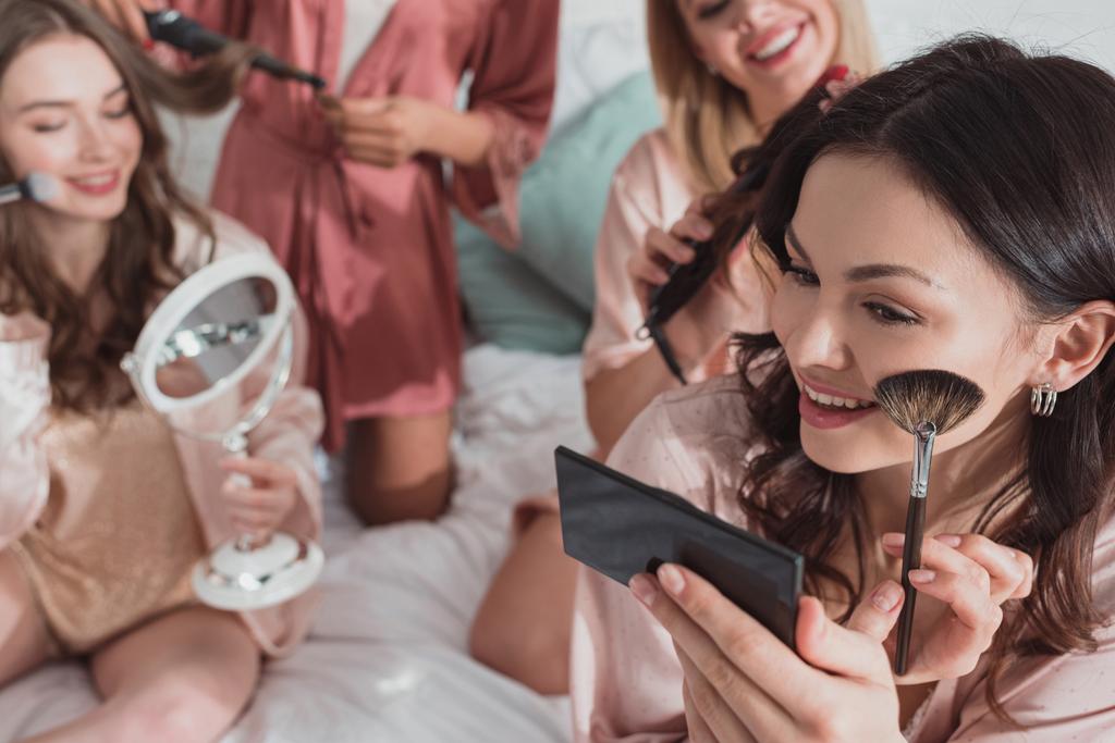 Καλλιεργημένη άποψη των πολυπολιτισμικών φίλων βάζοντας μακιγιάζ και κάνοντας χτενίσματα με σίδερα για μπούκλες στο κρεβάτι - Φωτογραφία, εικόνα