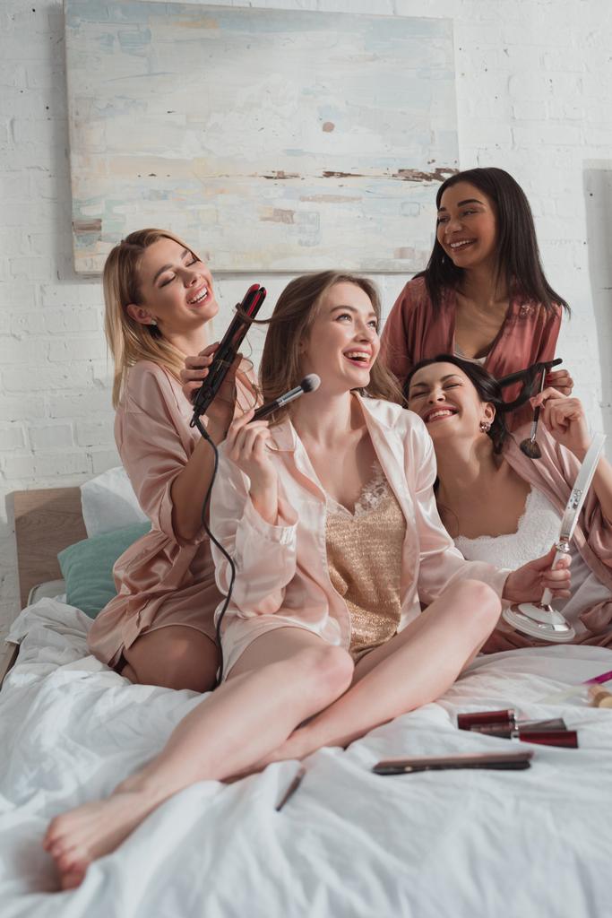 Selective focus of multiethnic women putting makeup and doing hairstyles at bachelorette party on bed - Photo, Image